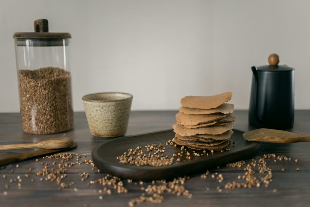 Oval plate with stack of healthy pancakes served on wooden table with glass jar of buckwheat grains and cup in kitchen